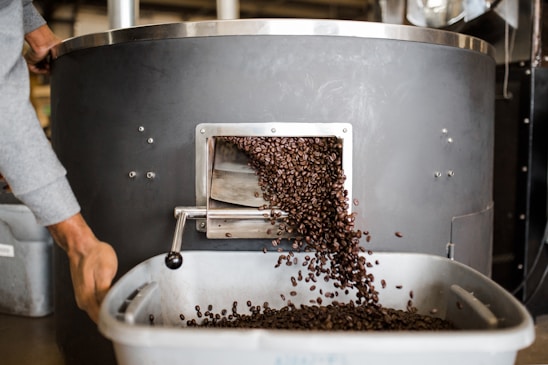 A large coffee roasting machine releases a stream of freshly roasted coffee beans into a nearby tray. A person is seen handling the equipment while beans cascade down. The setting appears to be a coffee roastery or industrial workspace.