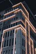 A modern building facade highlighted by bright, linear LED lights running along its edges and contours. The building has a geometric, structured design with vertical panels and numerous windows. The lighting creates a striking contrast against the dark night sky, emphasizing the building's architectural features.