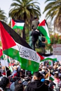 A large crowd gathers outdoors holding Palestinian flags with palm trees visible in the background. People are dressed in various clothing, and one individual is seen elevated above the crowd. Signs and traffic lights are also present in the scene, with vibrant colors and bustling activity.