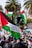 A large crowd gathers outdoors holding Palestinian flags with palm trees visible in the background. People are dressed in various clothing, and one individual is seen elevated above the crowd. Signs and traffic lights are also present in the scene, with vibrant colors and bustling activity.