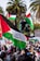 A large crowd gathers outdoors holding Palestinian flags with palm trees visible in the background. People are dressed in various clothing, and one individual is seen elevated above the crowd. Signs and traffic lights are also present in the scene, with vibrant colors and bustling activity.