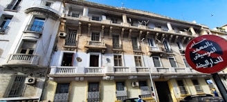 A multi-story building with a worn exterior, featuring balconies, ornate railings, and several satellite dishes. A red and black circular street sign with Arabic text is prominently displayed in front. The sky is clear and blue above.
