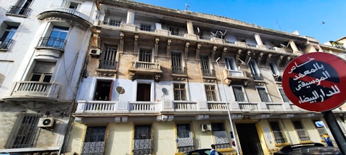 A multi-story building with a worn exterior, featuring balconies, ornate railings, and several satellite dishes. A red and black circular street sign with Arabic text is prominently displayed in front. The sky is clear and blue above.