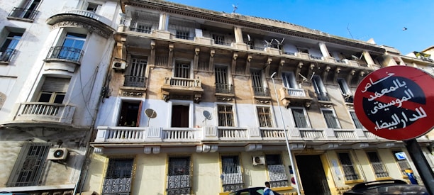 A multi-story building with a worn exterior, featuring balconies, ornate railings, and several satellite dishes. A red and black circular street sign with Arabic text is prominently displayed in front. The sky is clear and blue above.