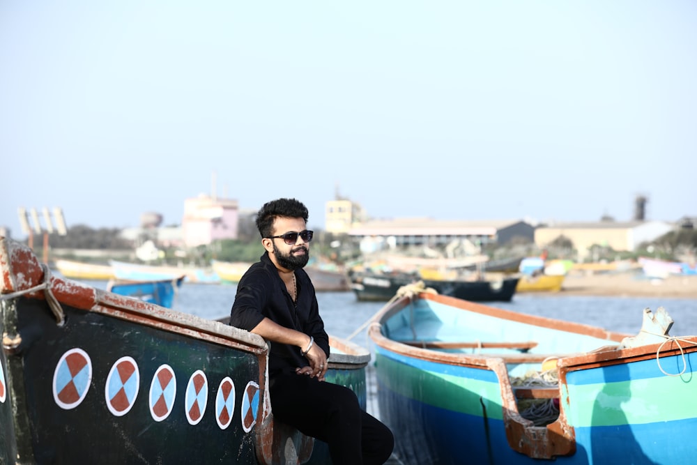 man in black jacket wearing black sunglasses standing beside blue and white boat during daytime