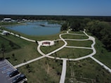 The photo showcases a well-manicured park with a large pond in the center. The pond has a wooden pier extending into it. Surrounding the pond are green lawns with a network of white walking paths. There is a circular seating area with a red centerpiece. Several cars are parked in a parking lot on the left side of the image, and a fence surrounds part of the park. The park is bordered by dense trees, and the sky above is clear and blue.