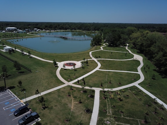 The photo showcases a well-manicured park with a large pond in the center. The pond has a wooden pier extending into it. Surrounding the pond are green lawns with a network of white walking paths. There is a circular seating area with a red centerpiece. Several cars are parked in a parking lot on the left side of the image, and a fence surrounds part of the park. The park is bordered by dense trees, and the sky above is clear and blue.
