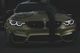A stylish and aggressive-looking BMW car with its headlights on, displaying bold text 'FAKE TAXI' on the bumper. The car features a sleek, shiny surface with a dark green stripe running down the center of the hood and is set against a dimly lit background.