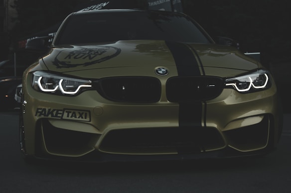 A stylish and aggressive-looking BMW car with its headlights on, displaying bold text 'FAKE TAXI' on the bumper. The car features a sleek, shiny surface with a dark green stripe running down the center of the hood and is set against a dimly lit background.