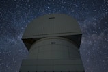 A large observatory structure with a domed top is silhouetted against a starry night sky. The structure appears industrial and robust, with panels and a geometric design. The night sky is filled with stars, creating a mesmerizing backdrop of cosmic beauty.