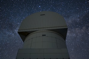 A large observatory structure with a domed top is silhouetted against a starry night sky. The structure appears industrial and robust, with panels and a geometric design. The night sky is filled with stars, creating a mesmerizing backdrop of cosmic beauty.