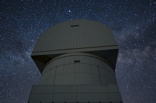 A large observatory structure with a domed top is silhouetted against a starry night sky. The structure appears industrial and robust, with panels and a geometric design. The night sky is filled with stars, creating a mesmerizing backdrop of cosmic beauty.