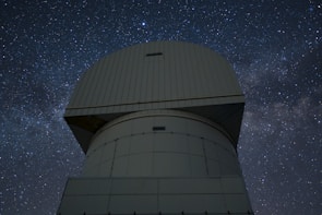 A large observatory structure with a domed top is silhouetted against a starry night sky. The structure appears industrial and robust, with panels and a geometric design. The night sky is filled with stars, creating a mesmerizing backdrop of cosmic beauty.
