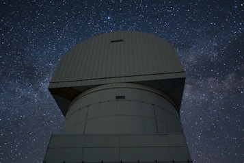 A large observatory structure with a domed top is silhouetted against a starry night sky. The structure appears industrial and robust, with panels and a geometric design. The night sky is filled with stars, creating a mesmerizing backdrop of cosmic beauty.