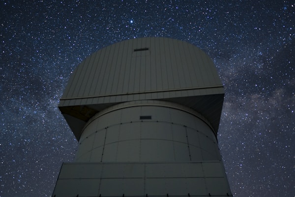 A large observatory structure with a domed top is silhouetted against a starry night sky. The structure appears industrial and robust, with panels and a geometric design. The night sky is filled with stars, creating a mesmerizing backdrop of cosmic beauty.