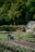 A lush garden scene with a wheelbarrow in the foreground, which contains gardening tools. In the background, there is a small shed and a variety of green foliage, including a flowering bush and a few small trees. The garden appears well-maintained with freshly tilled soil and neatly arranged plants.