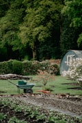 A lush garden scene with a wheelbarrow in the foreground, which contains gardening tools. In the background, there is a small shed and a variety of green foliage, including a flowering bush and a few small trees. The garden appears well-maintained with freshly tilled soil and neatly arranged plants.
