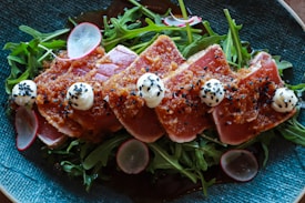 Slices of seared tuna are artfully arranged on a bed of fresh arugula. The tuna is topped with a flavorful seasoning and garnished with dollops of cream and black sesame seeds. Thinly sliced radishes add a splash of color.