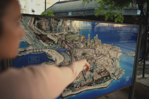 A person is pointing at a detailed illustrated map of Cádiz, featuring prominent landmarks and streets. The map is mounted on a stand in an outdoor setting, and the background shows a blurred view of the surrounding area with some greenery.