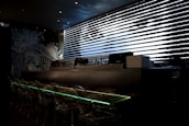 A dark, modern interior with a sushi bar featuring transparent seating and illuminated countertops. Black and white artistic wall designs create an abstract, oceanic theme. A sleek, horizontal window with patterned blinds adds a futuristic touch.