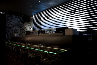 A dark, modern interior with a sushi bar featuring transparent seating and illuminated countertops. Black and white artistic wall designs create an abstract, oceanic theme. A sleek, horizontal window with patterned blinds adds a futuristic touch.