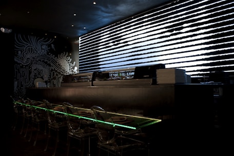 A dark, modern interior with a sushi bar featuring transparent seating and illuminated countertops. Black and white artistic wall designs create an abstract, oceanic theme. A sleek, horizontal window with patterned blinds adds a futuristic touch.