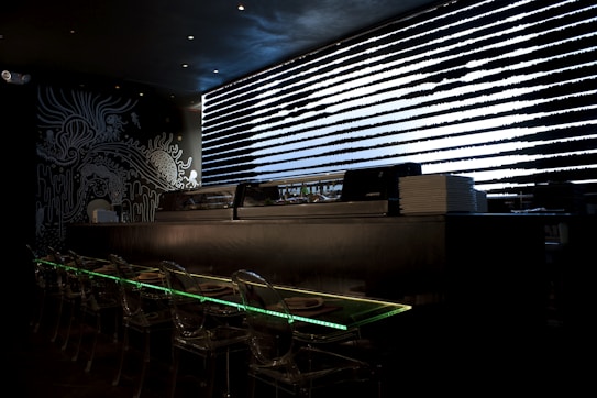 A dark, modern interior with a sushi bar featuring transparent seating and illuminated countertops. Black and white artistic wall designs create an abstract, oceanic theme. A sleek, horizontal window with patterned blinds adds a futuristic touch.