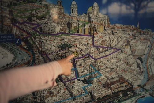 A detailed, colorful map of a city, prominently featuring various routes highlighted in purple and blue. Architectural drawings of significant landmarks and buildings are visible, with detailed illustrations suggesting it might be a tourist map. A hand is pointing toward a specific location on the map, indicating a focus or interest on that spot.