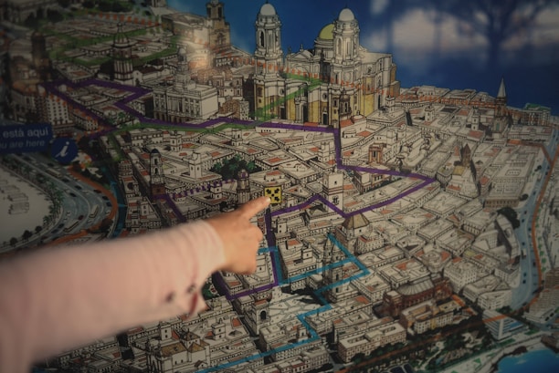 A detailed, colorful map of a city, prominently featuring various routes highlighted in purple and blue. Architectural drawings of significant landmarks and buildings are visible, with detailed illustrations suggesting it might be a tourist map. A hand is pointing toward a specific location on the map, indicating a focus or interest on that spot.