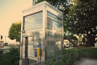 A metallic, glass-walled elevator shaft is surrounded by greenery and urban elements. Trees and plants provide a natural border around the structure, with a park-like setting evident in the background. A yellow control panel is mounted on the elevator exterior. A statue and pedestrians are visible in the distance, adding character to the open area.