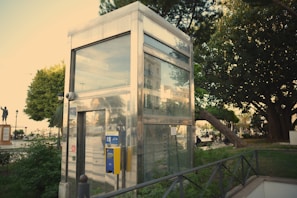 A metallic, glass-walled elevator shaft is surrounded by greenery and urban elements. Trees and plants provide a natural border around the structure, with a park-like setting evident in the background. A yellow control panel is mounted on the elevator exterior. A statue and pedestrians are visible in the distance, adding character to the open area.