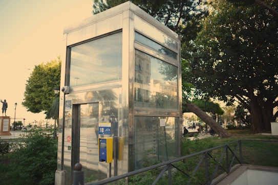A metallic, glass-walled elevator shaft is surrounded by greenery and urban elements. Trees and plants provide a natural border around the structure, with a park-like setting evident in the background. A yellow control panel is mounted on the elevator exterior. A statue and pedestrians are visible in the distance, adding character to the open area.