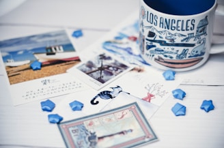 A Los Angeles-themed mug is placed on a white wooden surface surrounded by various colorful postcards and small blue paper stars. The postcards display scenic images and some contain foreign text. A subtle blue chameleon toy is also present among the items.