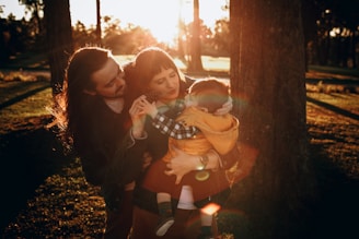 A family stands in a park during sunset, surrounded by trees. The man with long hair leans in close to a woman holding a small child dressed in a yellow jacket. Natural light creates a warm glow around them, highlighting the greenery and creating long shadows on the ground.