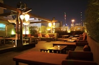 Wide shot of the rooftop lounge at night, city skyline glowing beyond the illuminated tables and shisha setups.