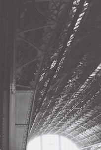 An intricately designed steel structure with riveted beams and a curved roof pattern. The interplay of light and shadow highlights the geometric complexity of the architecture, creating a dynamic visual effect.
