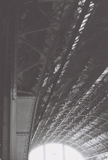 An intricately designed steel structure with riveted beams and a curved roof pattern. The interplay of light and shadow highlights the geometric complexity of the architecture, creating a dynamic visual effect.