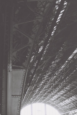 An intricately designed steel structure with riveted beams and a curved roof pattern. The interplay of light and shadow highlights the geometric complexity of the architecture, creating a dynamic visual effect.