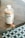 A bottle of a product labeled 'hella co.' and 'milktea hair therapy' is placed on a wooden surface. The bottle is round with a white cap and a minimalistic design featuring bubble tea graphics. The background includes a geometric floor pattern with circular motifs.