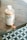 A bottle of a product labeled 'hella co.' and 'milktea hair therapy' is placed on a wooden surface. The bottle is round with a white cap and a minimalistic design featuring bubble tea graphics. The background includes a geometric floor pattern with circular motifs.