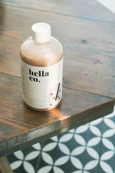 A bottle of a product labeled 'hella co.' and 'milktea hair therapy' is placed on a wooden surface. The bottle is round with a white cap and a minimalistic design featuring bubble tea graphics. The background includes a geometric floor pattern with circular motifs.