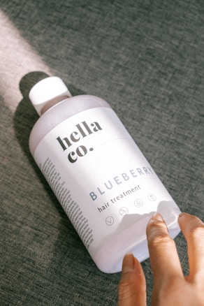 A bottle of hair treatment labeled 'hella co. BLUEBERRY hair treatment' rests on a textured gray surface. Sunlight casts a soft glow on the bottle, with the shadow clearly visible. A hand is reaching towards the bottle, suggesting interaction.
