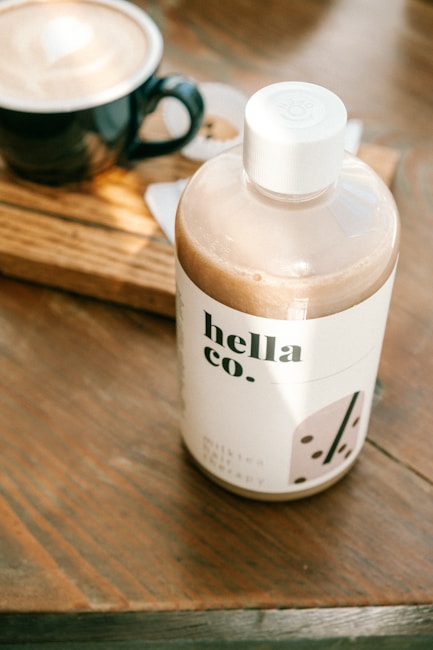 A bottle labeled 'hella co.' filled with a light brown liquid is placed on a wooden surface next to a black cup filled with a latte. The label on the bottle has minimalistic graphics and text in black. The table shows a warm, natural wood grain texture, and sunlight is casting soft shadows across the scene.