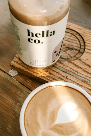 A bottle labeled 'hella co. milk tea hair therapy' rests on a wooden coaster. In the foreground, there is a cup of coffee with a heart-shaped latte art. The setting is on a wooden table with a warm ambiance.