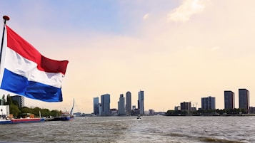 A Dutch flag waves prominently in the foreground, with a city skyline dominated by modern high-rise buildings in the background. The image includes the iconic Erasmus Bridge spanning across the river. The water in the foreground has a rippled surface, reflecting the soft, warm light of the sky.