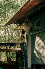 A partially open roller shutter attached to a building with blue walls. Above it, an ornate chandelier hangs under a canopy. A nearby tree creates dappled sunlight patterns on the surfaces. Signs with directions are visible, possibly indicating a pathway. Cables are visible running along the walls.