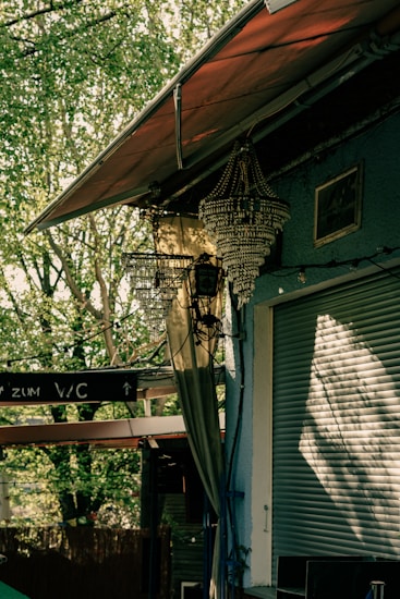 A partially open roller shutter attached to a building with blue walls. Above it, an ornate chandelier hangs under a canopy. A nearby tree creates dappled sunlight patterns on the surfaces. Signs with directions are visible, possibly indicating a pathway. Cables are visible running along the walls.