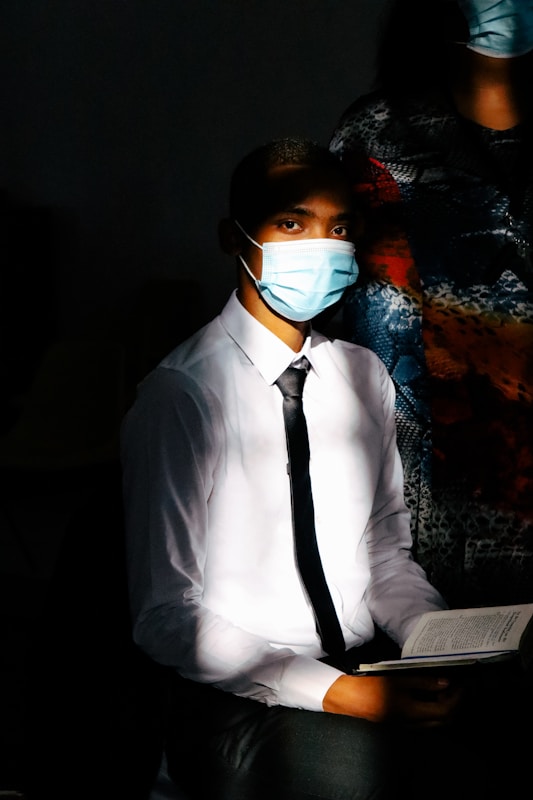 A person wearing a white shirt, black tie, and medical mask is sitting in a dimly lit room. The individual is holding an open book, with sunlight partially illuminating the face and shirt, creating a strong contrast with the dark background.
