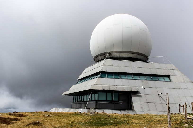 A large radar dome structure sits atop a multi-tiered building. The dome is situated against a backdrop of overcast skies, with grassy earth in the foreground.