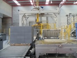 An industrial setting featuring a large machinery setup for handling cinder blocks. The foreground shows a stack of gray cinder blocks neatly arranged on a wooden pallet. Yellow safety railings and metal components are prominently visible. The area is well-lit by overhead lights and a structured metal framework forms the ceiling.